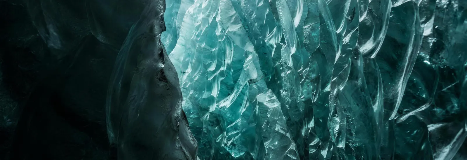 The crystalline teal wall of a glacier crevasse, angular fracture planes lit from within the ice