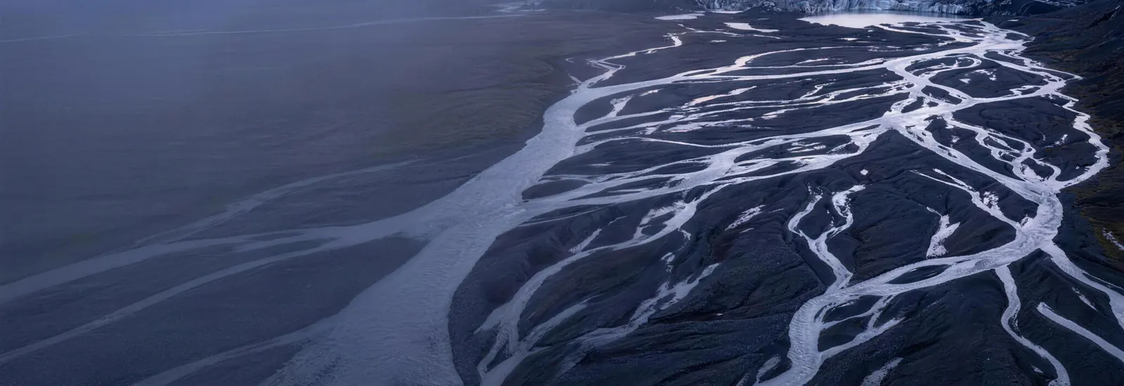 A braided glacial river delta at dawn, dozens of silver meltwater channels weaving across dark gravel flats